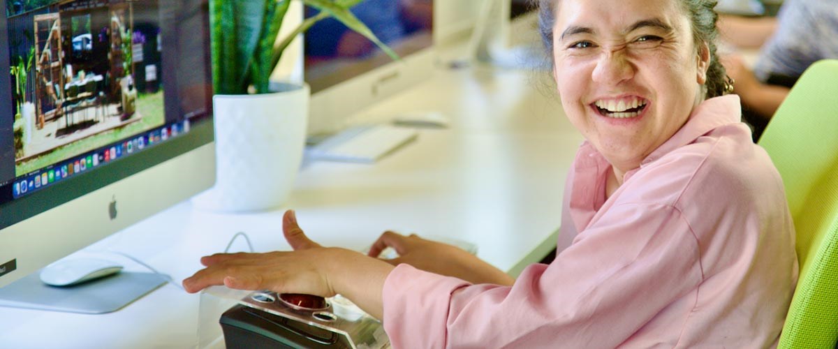 Bella, wearing a pink blouse, happily works at a computer using a Kensington trackball.