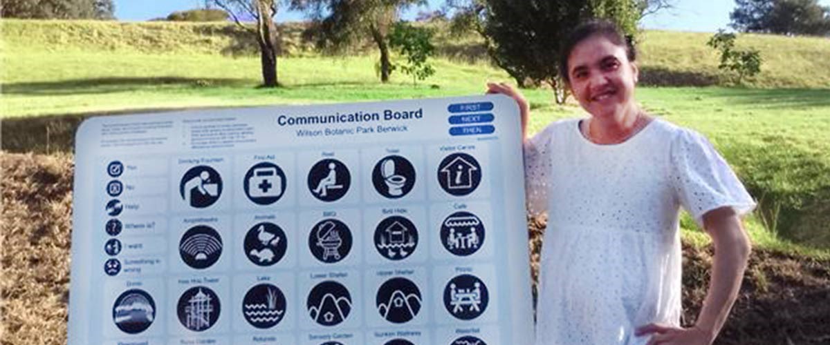Bella Firth, a young woman in a white dress, smiles while pointing to an accessible communication board at Wilson Botanic Park.