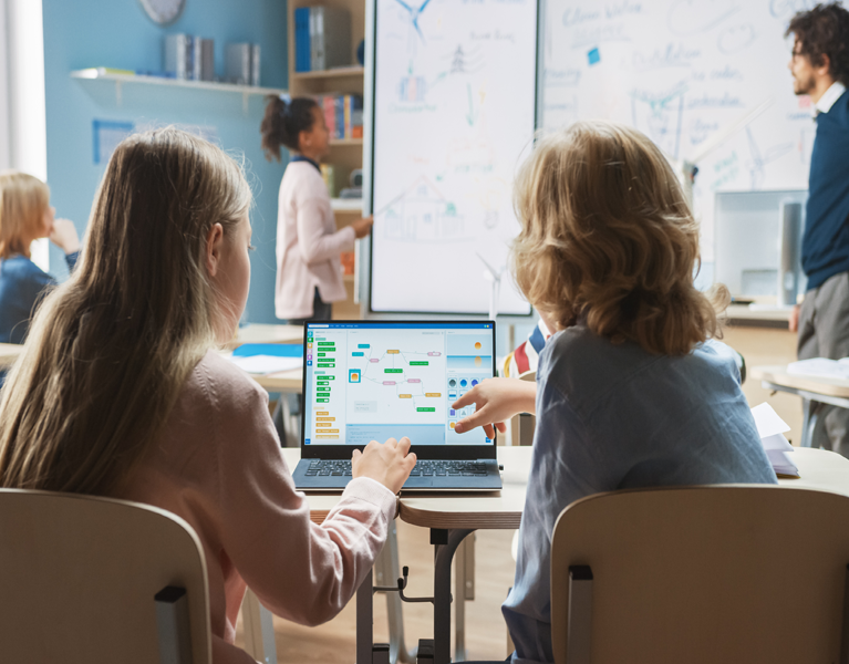 2 children working on a shared laptop with a teacher at the front of the classroom.