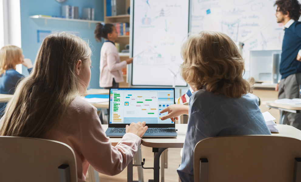 2 children working on a shared laptop with a teacher at the front of the classroom.