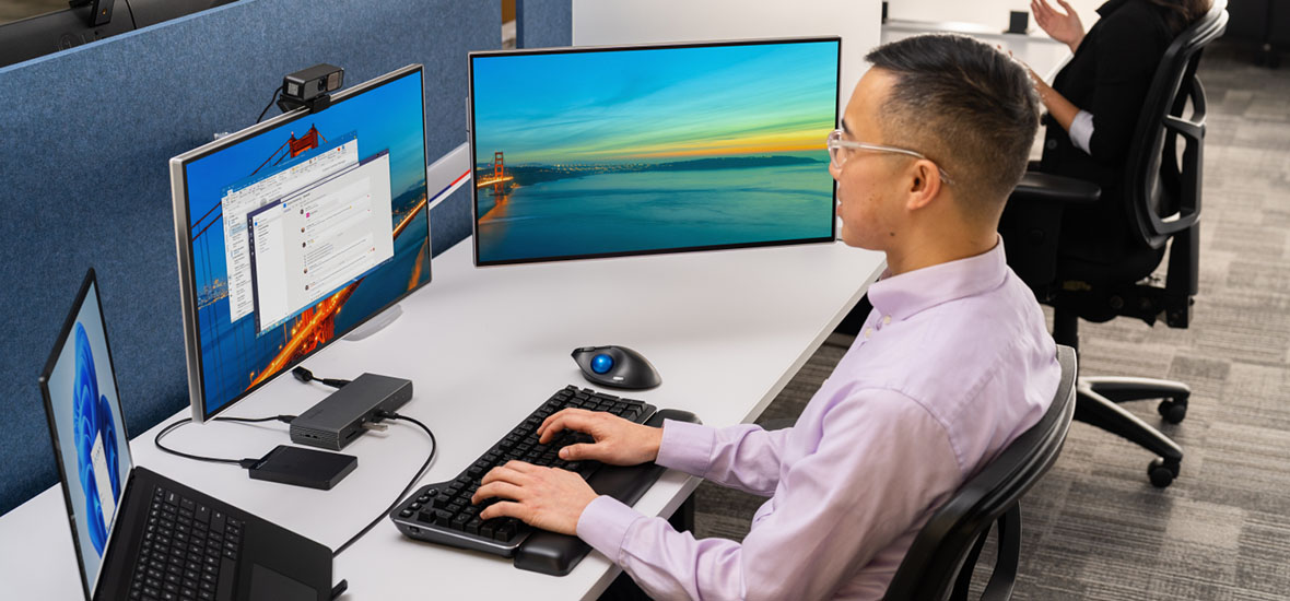 Office worker using a dual-monitor setup connected to a docking station, ergonomic keyboard, and trackball mouse at a modern workstation, with another colleague nearby.
